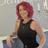 A warm, confident woman with softly curled dark hair smiles warmly in a beautifully lit salon. She’s wearing a silver-grey top and heart-shaped pendant, standing near a glowing candelabra against a backdrop that reads “beautiful.” The setting feels calm, elegant, and welcoming – just like her.
