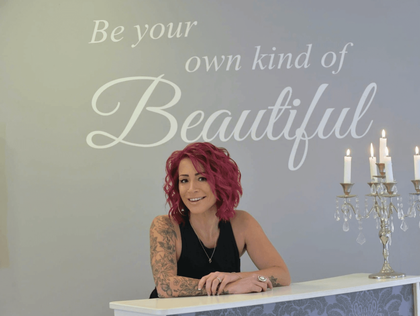 Belle, the salon owner, standing behind the reception desk with a warm smile, in front of a wall with the quote “Be your own kind of Beautiful”.