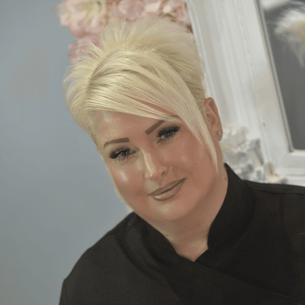 Donna, a professional beauty therapist and hair stylist at The Dolls House in Eastbourne, smiling warmly in front of a salon mirror and floral background.