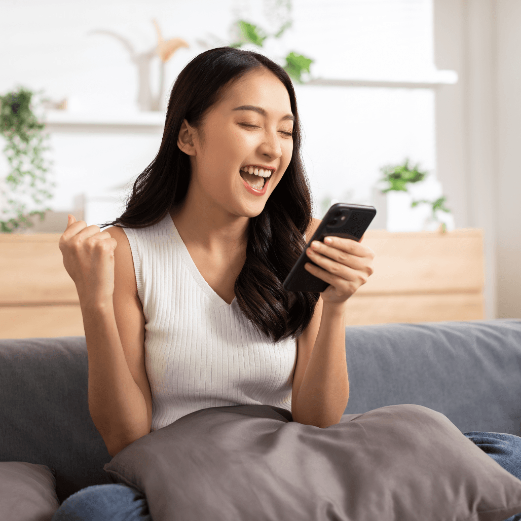 Excited young woman smiling while looking at her phone, celebrating a surprise message – perfect for illustrating the joy of receiving a Dolls House Hair & Beauty Boutique e-gift card.