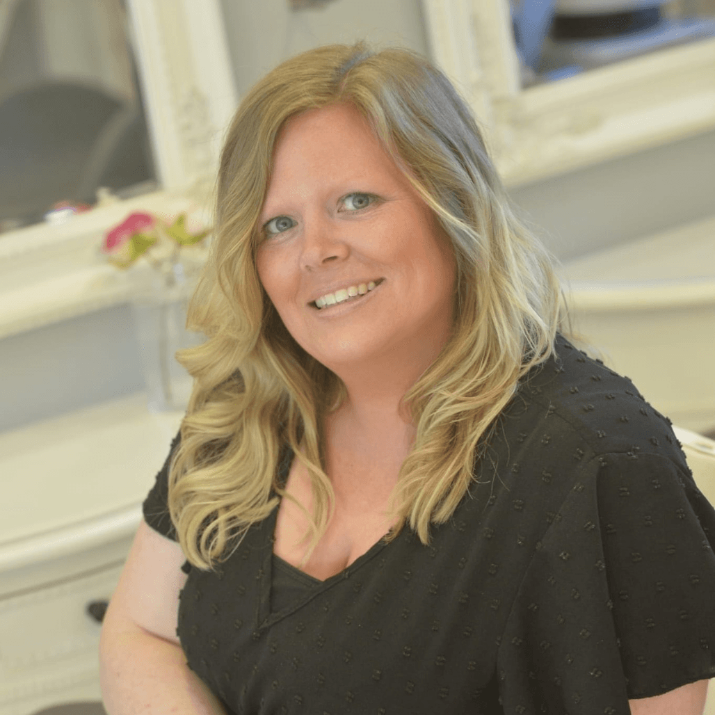 Stacey, a friendly and professional hair stylist at The Dolls House in Eastbourne, smiling warmly in the salon with soft waves and a relaxed black top.